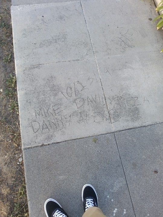 Names scratched into a concrete sidewalk: 10/13/14, Mike, Danny, David, Alexs, Reza, with a pair of black Vans at the bottom of the frame