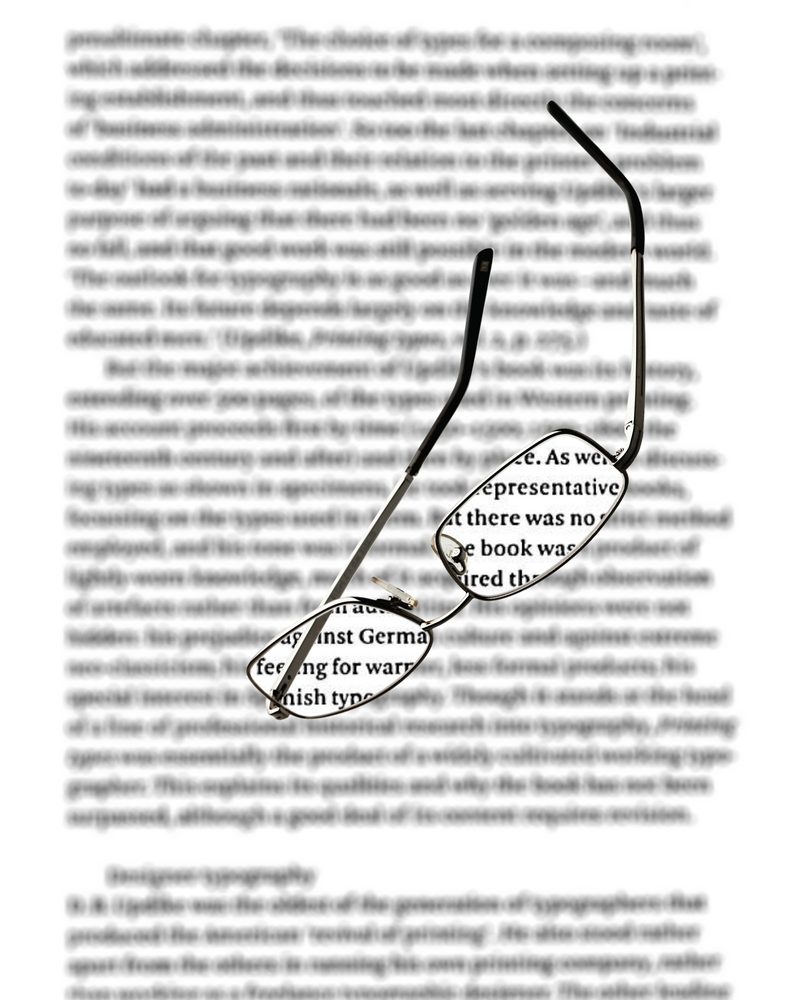 A page of Modern Typography blurred as someone who needs +2.25 reading glasses sees it without them, with reading glasses lying on the page; through the lenses the text is sharp and slightly larger